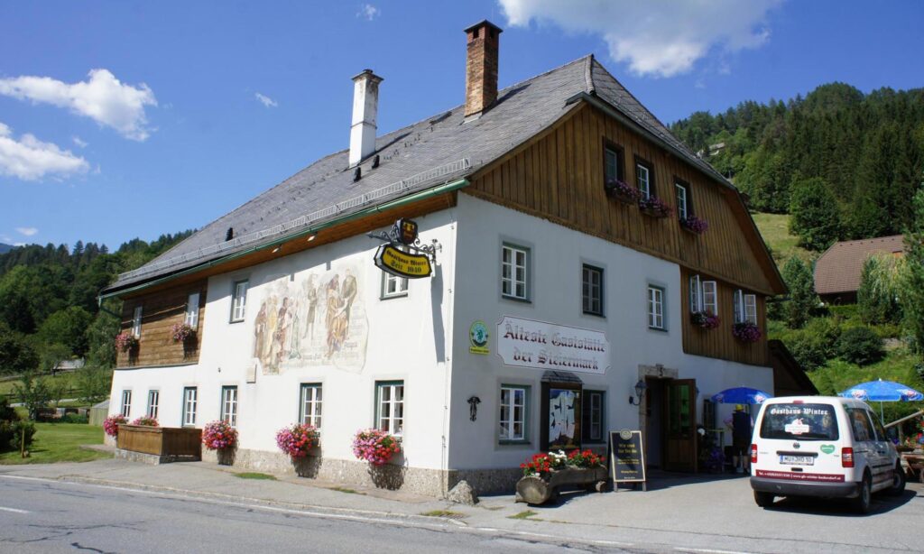 Außenansicht des historischen Gasthauses Winter mit Blumenkästen, Holzelementen und Berglandschaft im Hintergrund.